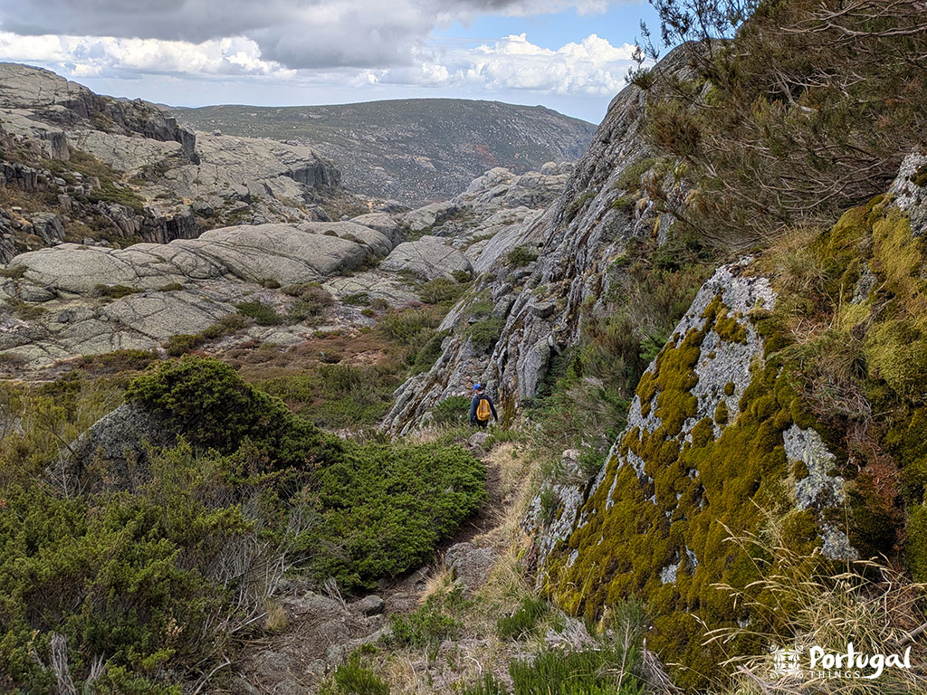 Uma pessoa com uma mochila caminha ao longo de um trilho desafiante estreito nas montanhas rochosas e cobertas de arbustos da Serra da Estrela; manchas de musgo e um céu parcialmente nublado acrescentam atmosfera. 
