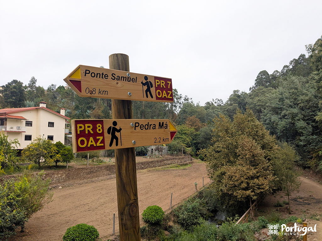 Wooden sign indicating the walking directions: 0.8 km to Ponte Samüel (PR 7 OAZ) on the left and 2.2 km to Pedra Má (PR 8 OAZ) on the Pindelo walkways, with trees, a house and a dirt path in the background.