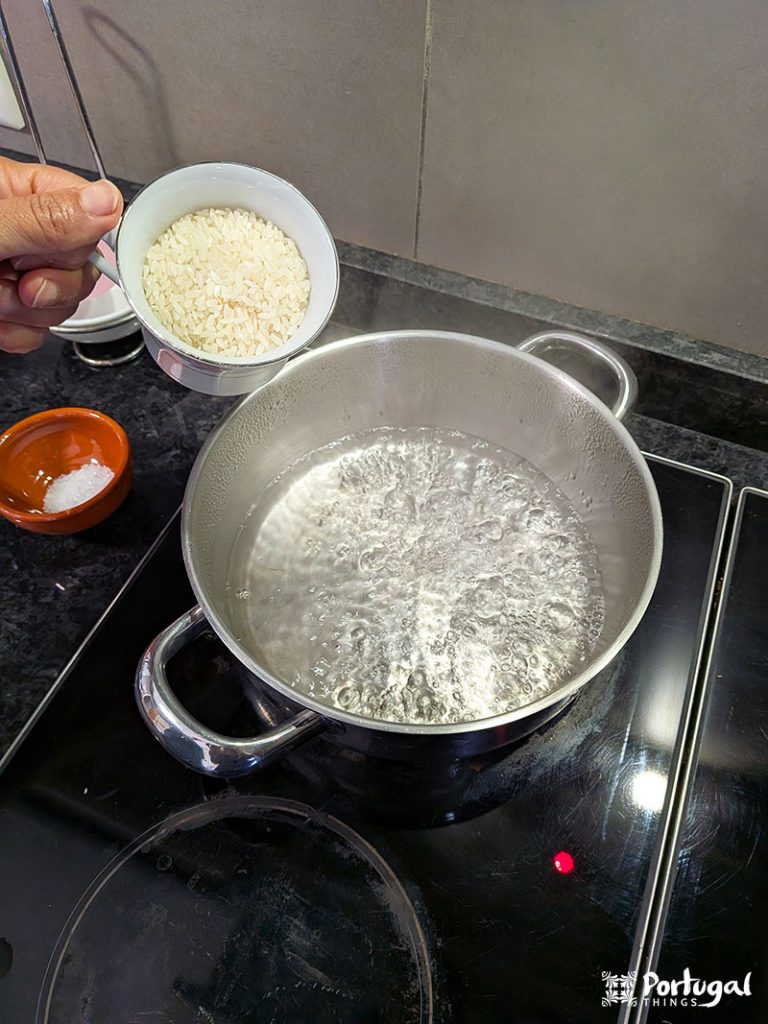 A hand holds a small bowl of uncooked white rice over a pot of boiling water on the stove, ready to begin a rice pudding recipe. A small brown dish of salt sits near the counter, against a tiled kitchen wall.