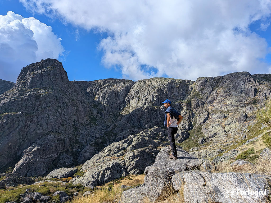 Uma pessoa com uma mochila e um boné azul está em um afloramento rochoso ao longo do trilho desafiante da Serra da Estrela, admirando uma paisagem montanhosa acidentada sob um céu parcialmente nublado com vegetação esparsa.