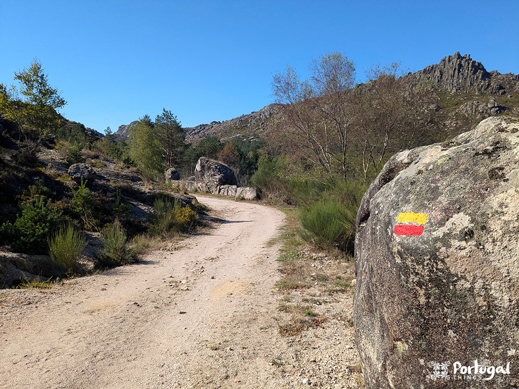 A dirt trail, part of the PR3 Castrejo Trail near Castro Laboreiro, winds through a rocky mountain landscape dotted with shrubs and trees. A large rock in the foreground displays a red and yellow trail marker under a clear blue sky.