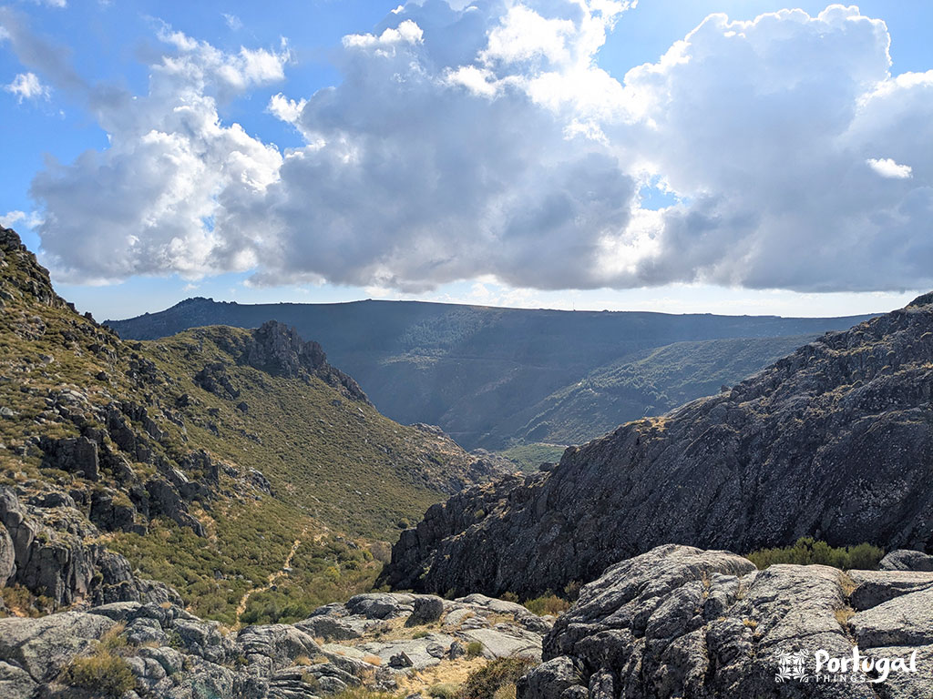 Uma paisagem de montanha rochosa com vegetação verde, penhascos escarpados e um caminho sinuoso sob um céu parcialmente nublado. A luz do sol atravessa as nuvens, iluminando partes da Rota do Maciço Central da Serra da Estrela. 