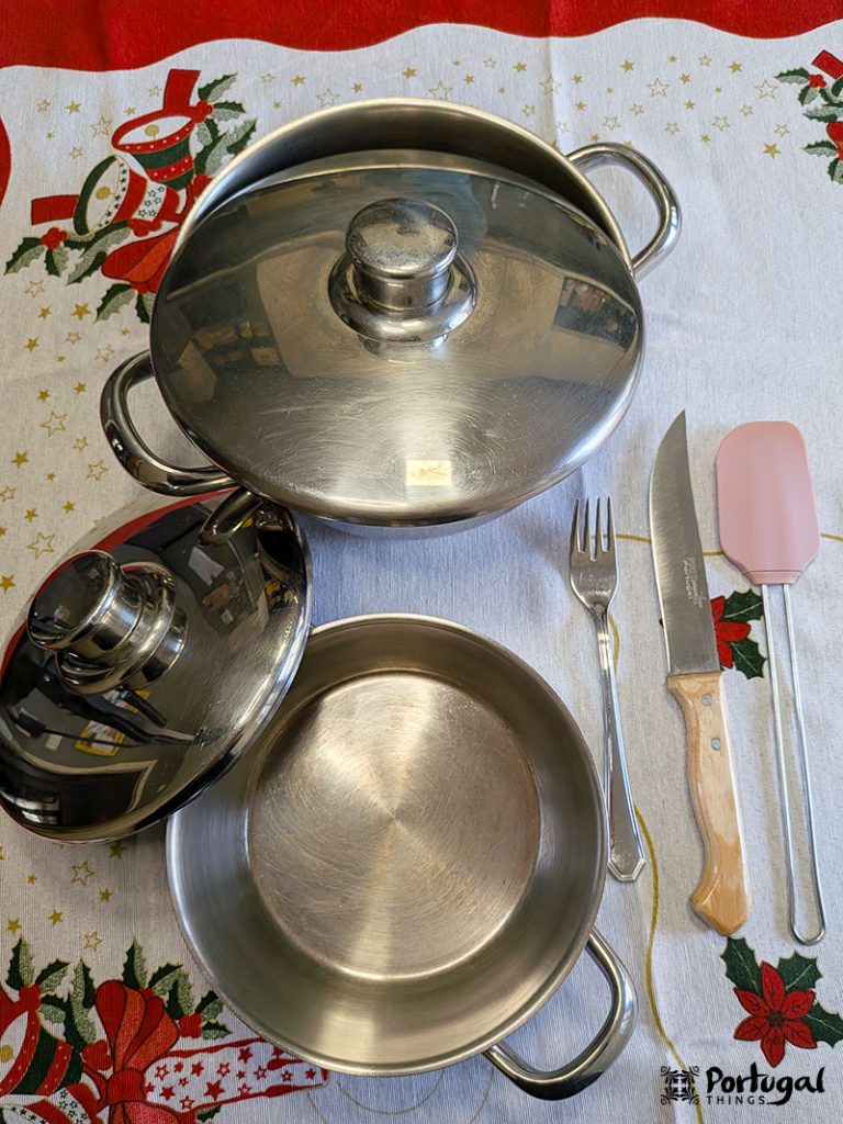 Two stainless steel pots with lids, a fork, a knife with a wooden handle and a pink spatula are arranged on a festive tablecloth decorated with festive motifs.