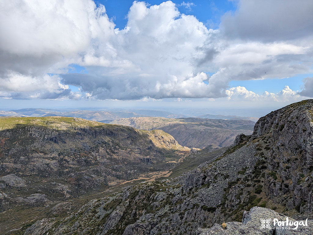 Paisagem de montanha rochosa sob um céu parcialmente nublado com manchas verdes, colinas distantes e sombras projetadas pelas nuvens. O terreno acidentado da Serra da Estrela define o cenário para este trilho desafiante