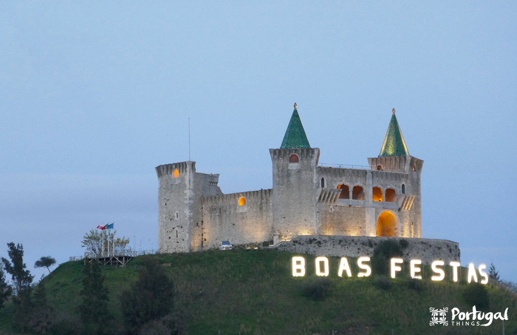 A stone castle with green-roofed towers sits on a hill at dusk, illuminated by lights. Large white letters on the hillside spell out 