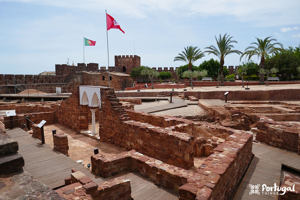 Stone ruins and preserved walls of Silves Castle, one of the most beautiful castles in Portugal, with two flags flying, palm trees and information plaques; the sky is partly cloudy.