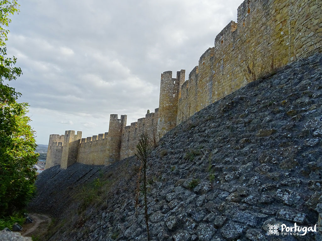 A stone wall of a medieval castle with several watchtowers rises along a rocky hillside under a cloudy sky. The vegetation is sparse near the wall, highlighting one of the most beautiful castles and a tourism icon in Portugal.