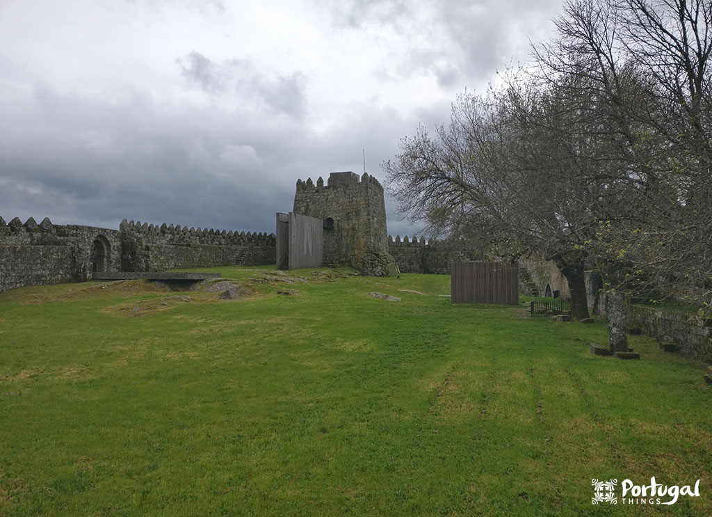 A medieval stone castle with crenellated walls and a round tower stands on a lawn under a cloudy sky. Leafless trees line the right side, showcasing one of the beautiful castles featured in Castles of Portugal.