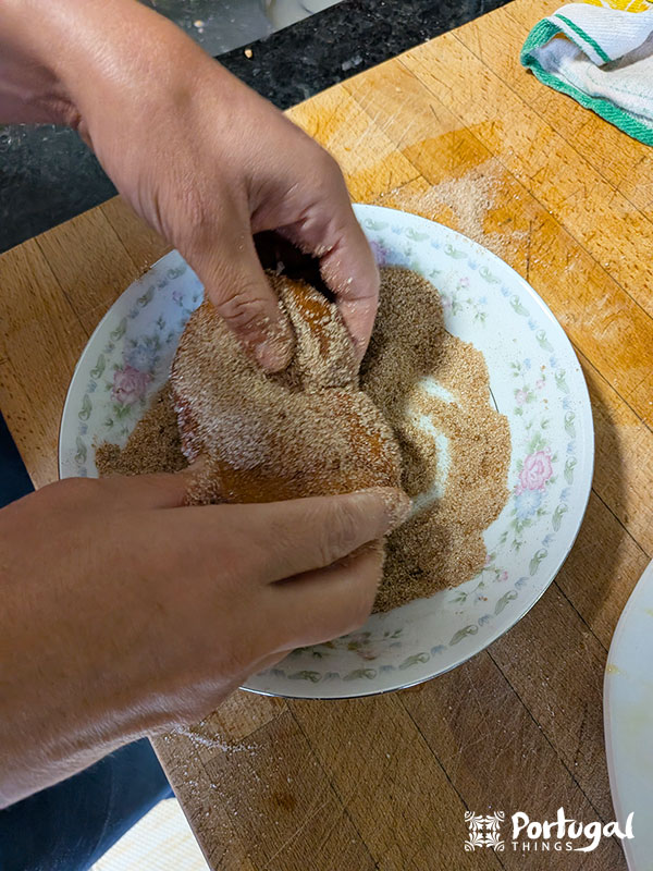 Duas mãos cobrem um pedaço de massa frita, conhecida como Rabanadas de Leite, com uma mistura de açúcar e canela em um prato com estampa floral, colocado em uma superfície de madeira. Uma toalha e parte da bancada indicam que essa receita tradicional está em andamento.