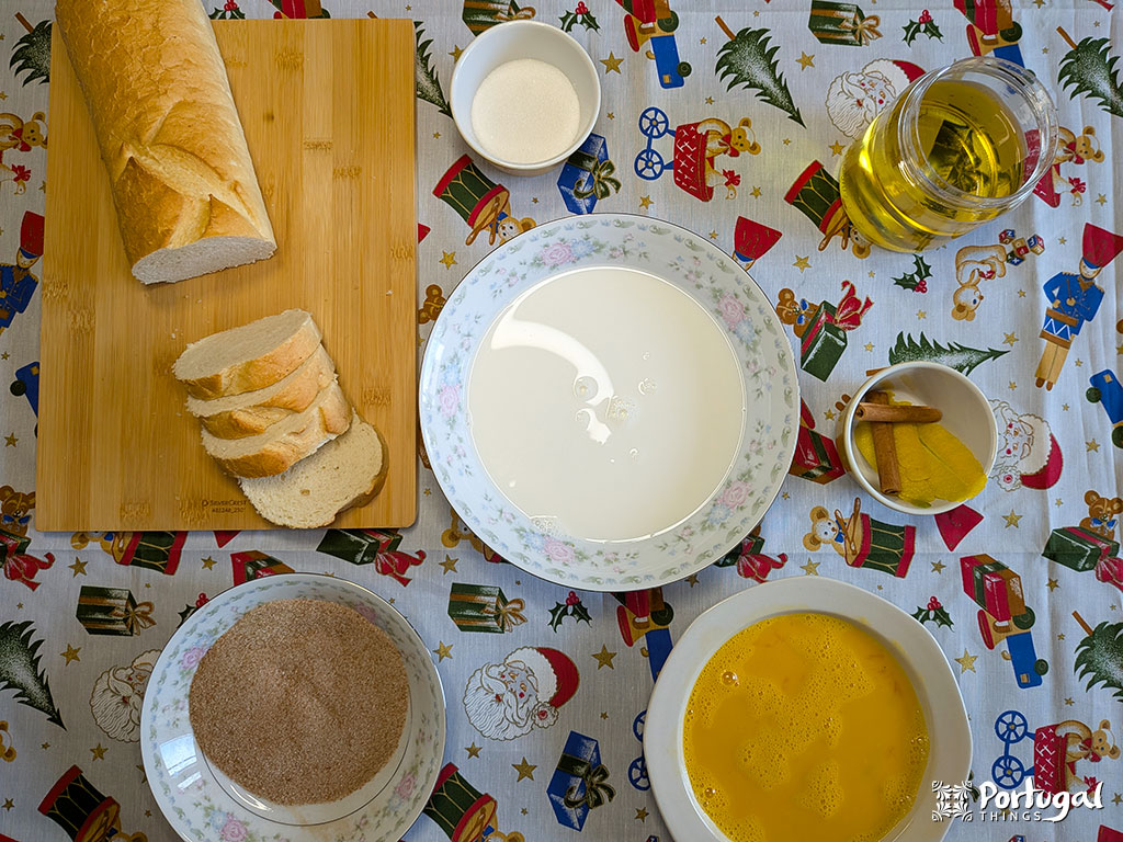 Uma mesa posta para Rabanadas de Leite: um pão fatiado em uma tábua de madeira, tigelas com ovos batidos, leite, açúcar, açúcar com canela e uma jarra de vidro com azeite de oliva em uma toalha de mesa com tema natalino - perfeito para esta receita tradicional.