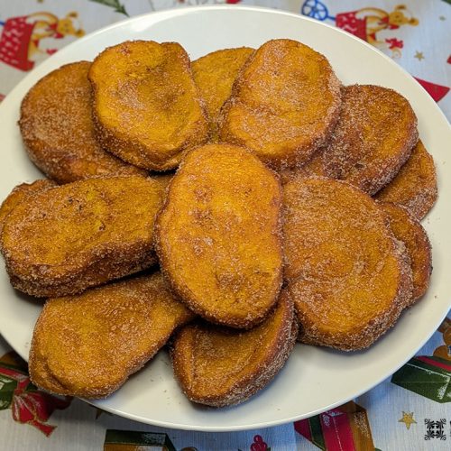 Um prato branco contém uma pilha de rabanadas de leite douradas e cobertas com açúcar. O prato é colocado em uma toalha de mesa festiva com padrões natalinos, refletindo essa receita tradicional apreciada durante as festas de fim de ano.