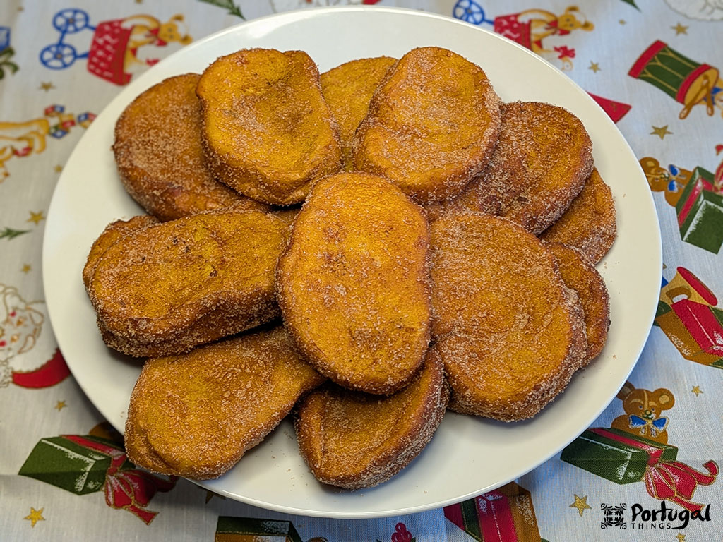 Um prato branco contém uma pilha de rabanadas de leite douradas e cobertas com açúcar. O prato é colocado em uma toalha de mesa festiva com padrões natalinos, refletindo essa receita tradicional apreciada durante as festas de fim de ano.