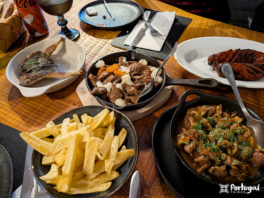 Uma mesa de madeira com pratos de batatas fritas, carnes grelhadas, linguiça e carne cozida, além de uma lata de refrigerante e pão - mostrando onde comer no Gerês para autênticos pratos portugueses.