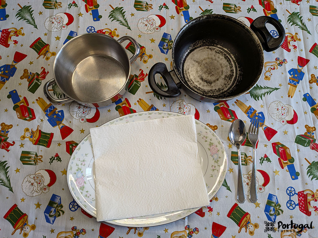 Uma mesa com uma toalha de mesa com tema natalino exibe uma panela de metal vazia, uma panela preta vazia e um conjunto de talheres - prontos para preparar rabanadas de leite, uma receita tradicional perfeita para as festas de fim de ano.
