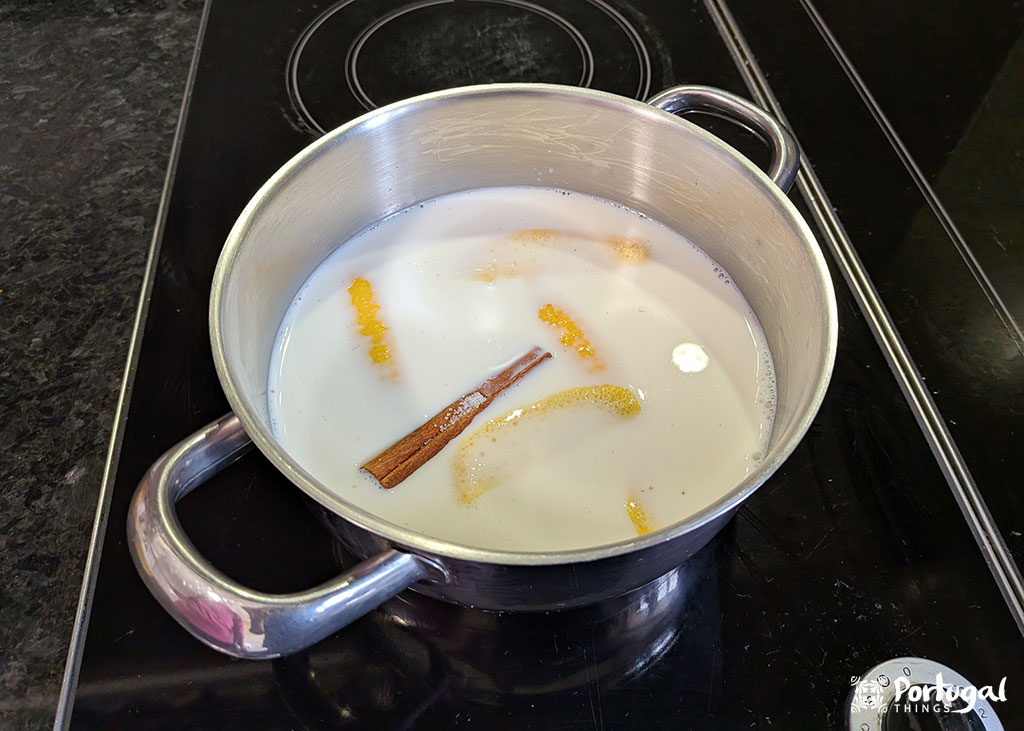 Uma panela de aço inoxidável em um fogão contém leite, paus de canela e tiras de casca de laranja - ingredientes essenciais para uma receita tradicional de rabanadas de leite - sendo aquecidos suavemente, com espuma e especiarias visíveis na superfície.