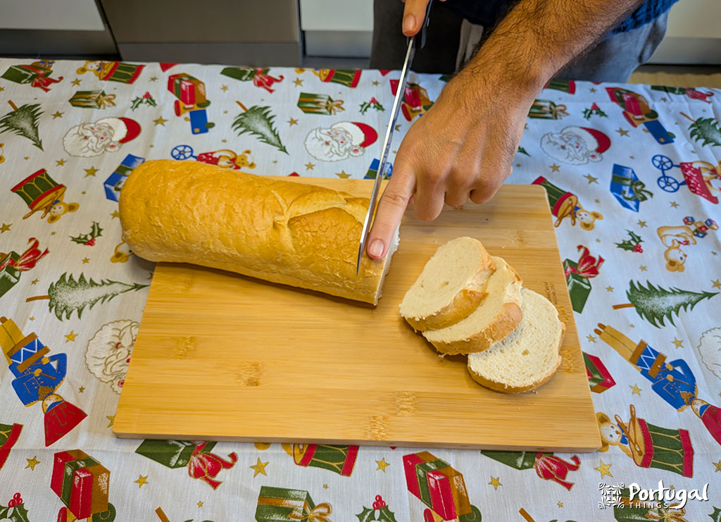 Uma pessoa corta um pão em uma tábua de corte de madeira, preparando Rabanadas de Leite - uma receita tradicional. A toalha de mesa festiva apresenta desenhos com temas natalinos