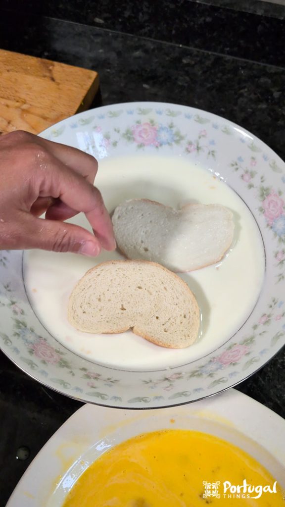 Uma mão mergulha fatias de pão em uma tigela rasa com padrão floral cheia de leite, preparando Rabanadas de Leite. Outra tigela contendo um líquido amarelo, provavelmente ovos batidos para essa receita tradicional, está parcialmente visível na parte inferior da imagem.