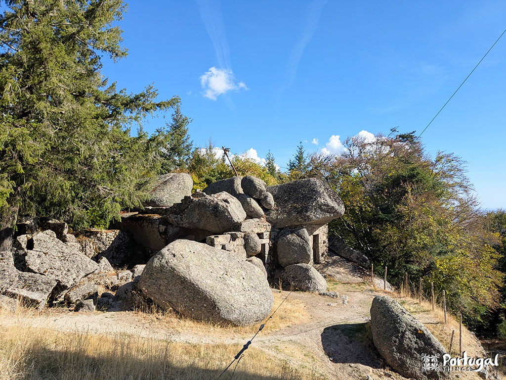 Large granite stones are stacked to form a rustic stone house in Penhas Douradas, Serra da Estrela, surrounded by trees and dry grass under a blue sky. A fence runs along the right side.