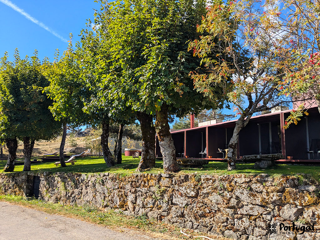 A row of leafy trees lines a stone wall in front of a single-story building with a red roof and shaded balcony, a typical scene along the Serra da Estrela trail near Penhas Douradas. The grass is green, and the sky is clear and blue.