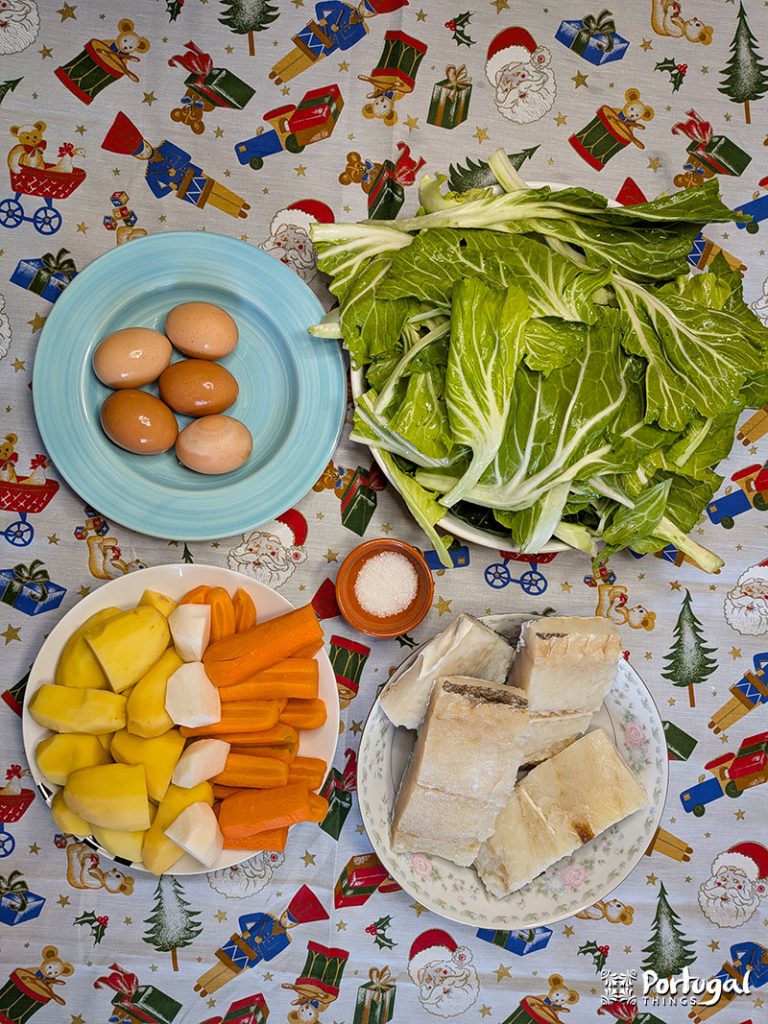 Top-down view of a festive table set for Bacalhau com Todos, with four brown eggs on a blue plate, leafy greens, a bowl of cut potatoes and carrots, salt, and white bread, all arranged on a Christmas-themed tablecloth.