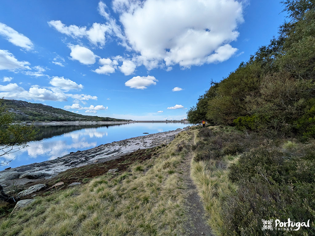 A dirt path, part of an easy trail, runs alongside a calm lake with grassy and rocky shores near Penhas Douradas in Serra da Estrela. Trees line the right side, and the sky is partly cloudy.