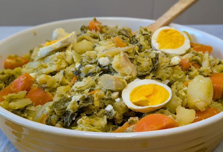 Uma tigela cheia de uma mistura de legumes picados, incluindo cenouras e verduras, coberta com fatias de ovo cozido. Uma colher de pau está descansando dentro da tigela - uma convidativa Receita de Natal inspirada no clássico Roupa Velha.