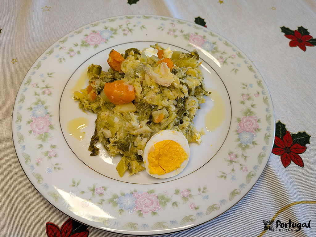 A plate of traditional Portuguese Roupa Velha, with shredded salted cod, potatoes, carrots, cabbage, and a slice of hard-boiled egg, served on a floral-patterned plate—a classic Christmas recipe on a festive tablecloth.