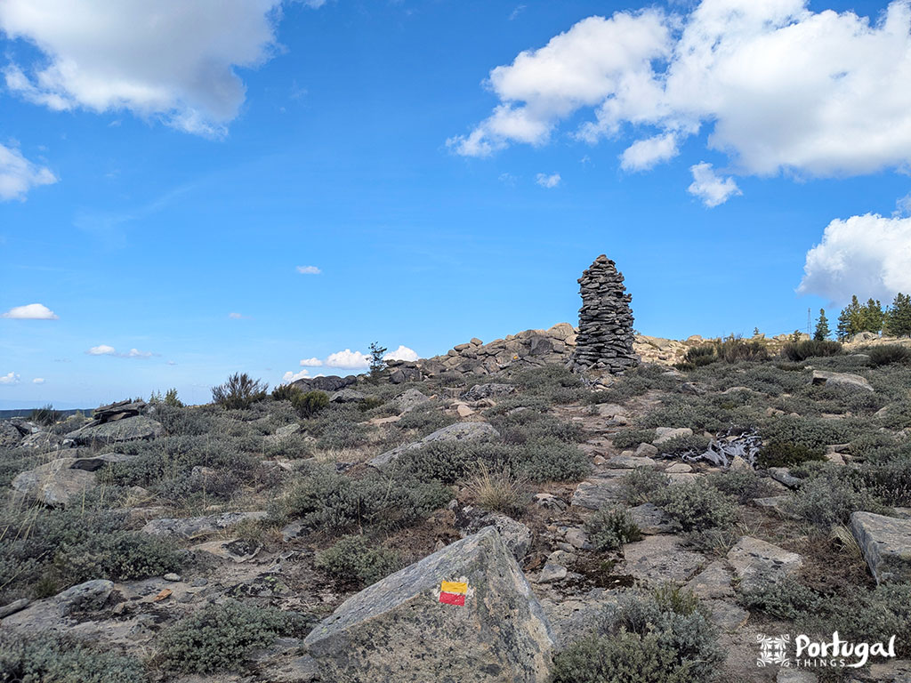 Rocky landscape with scattered low shrubs and a tall stone marker in the center, located along the Serra da Estrela trail near Penhas Douradas. A rock in the foreground displays a red and yellow trail marker.