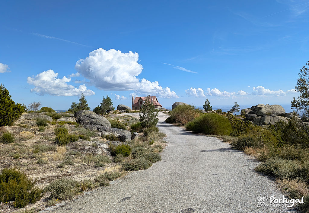 A paved path winds through rocky terrain dotted with shrubs and trees, leading to a house under a blue sky with large clouds, located in Penhas Douradas, Serra da Estrela.