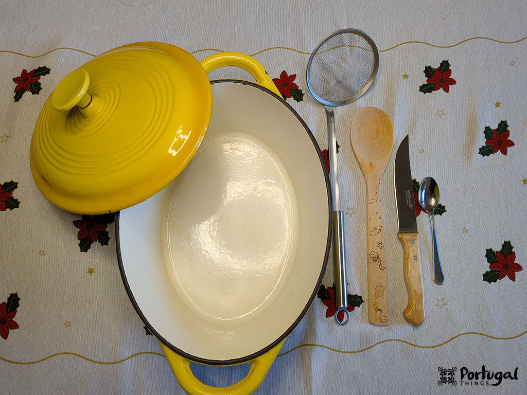 A large oval pot with a yellow lid sits open on a table with a floral tablecloth—perfect for preparing Roupa Velha, a classic Christmas recipe. Nearby are a strainer, wooden spoon, meat fork, and knife.