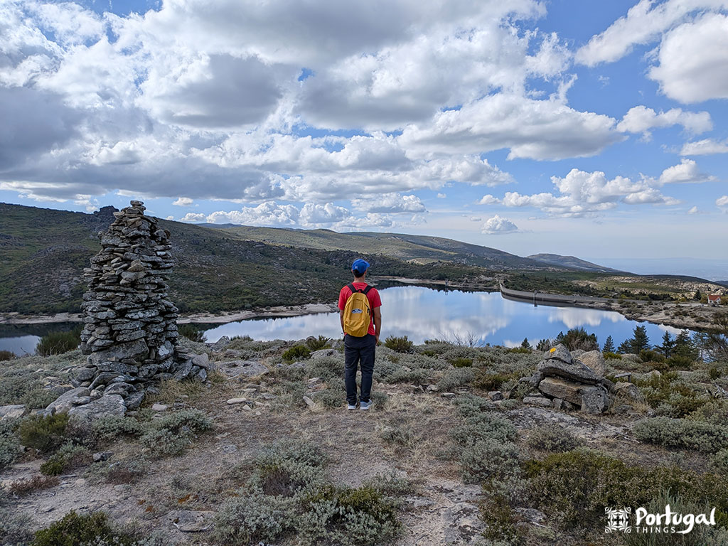 A person with a yellow backpack stands on rocky terrain in Serra da Estrela, overlooking a calm lake surrounded by hills and clouds. A stack of piled stones is to the left.