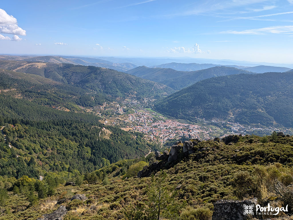 A panoramic view of a green valley and a small town near Serra da Estrela, with wooded hills and mountains under a blue sky. The words “Portugal Things” appear in the bottom right corner, highlighting this easy trail to Penhas Douradas.