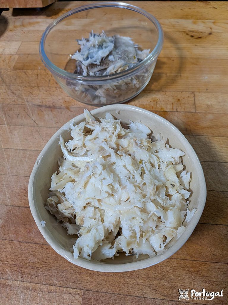 Shredded cod in a beige bowl on a wooden surface, with a smaller glass bowl containing fish skin and bones in the background.