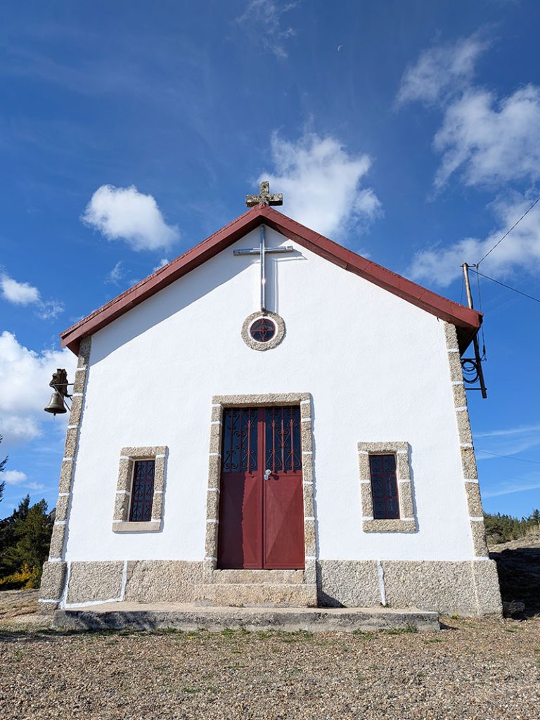 Small, charming white chapel with a red door and two barred windows, stone finishes, and a cross on the roof. Located along the Penhas Douradas Loop trail in Serra da Estrela. A metal bell hangs to the left, with scattered clouds dotting the blue sky.