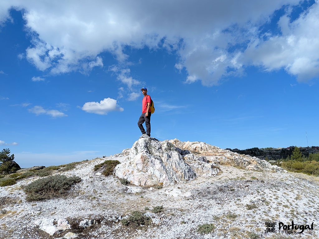 A person wearing a red shirt and a backpack stands atop a rocky hill under a blue sky with scattered clouds in Penhas Douradas, Serra da Estrela. Sparse vegetation surrounds the rocks.