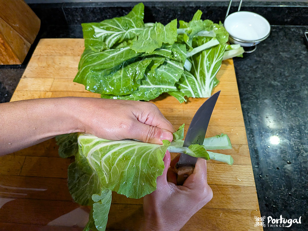 A person uses a knife to cut the thick stem of a leafy green vegetable on a wooden cutting board, preparing ingredients for a codfish recipe. More chopped greens are in the background, with a small white bowl nearby.