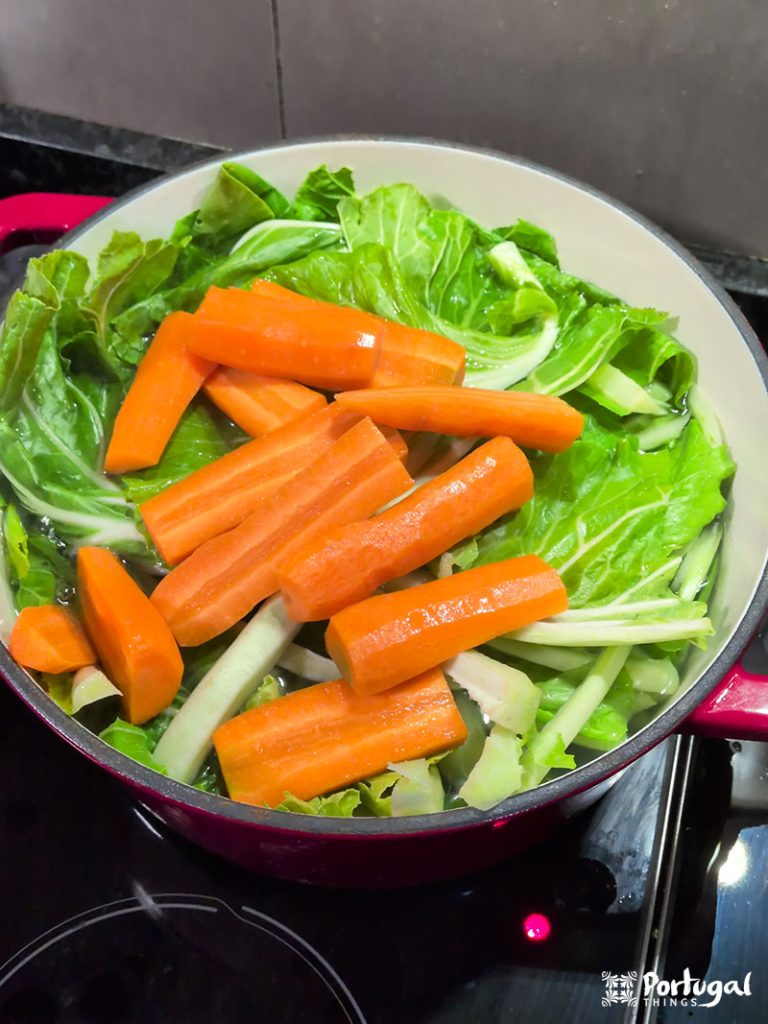 A pot on the stove filled with water, cabbage leaves, and several peeled and chopped carrots—vegetables cooking as part of a traditional Bacalhau com Todos recipe.