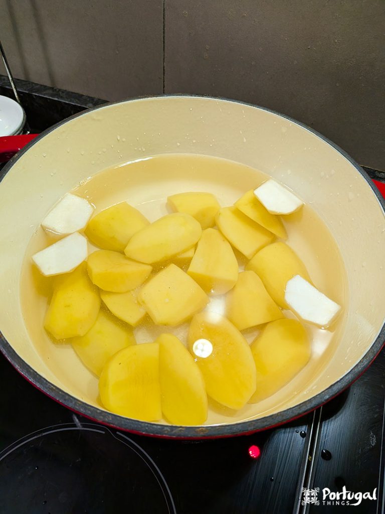 A pot filled with peeled and chopped potatoes and a few pieces of white fat in the water sits on the stove—just the beginning of a comforting recipe like Bacalhau com Todos, ready to be cooked to perfection.