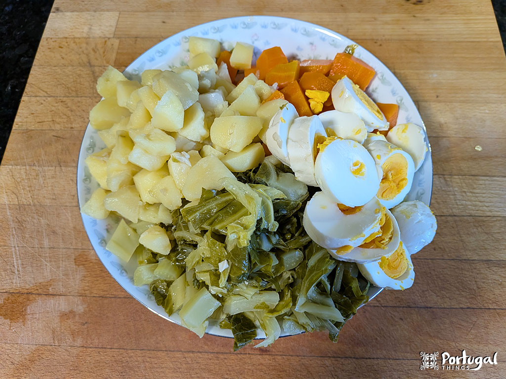 A plate of Roupa Velha, a traditional Christmas recipe, featuring boiled potatoes, sliced hard-boiled eggs, cooked carrots, and greens arranged in sections on a wooden cutting board.