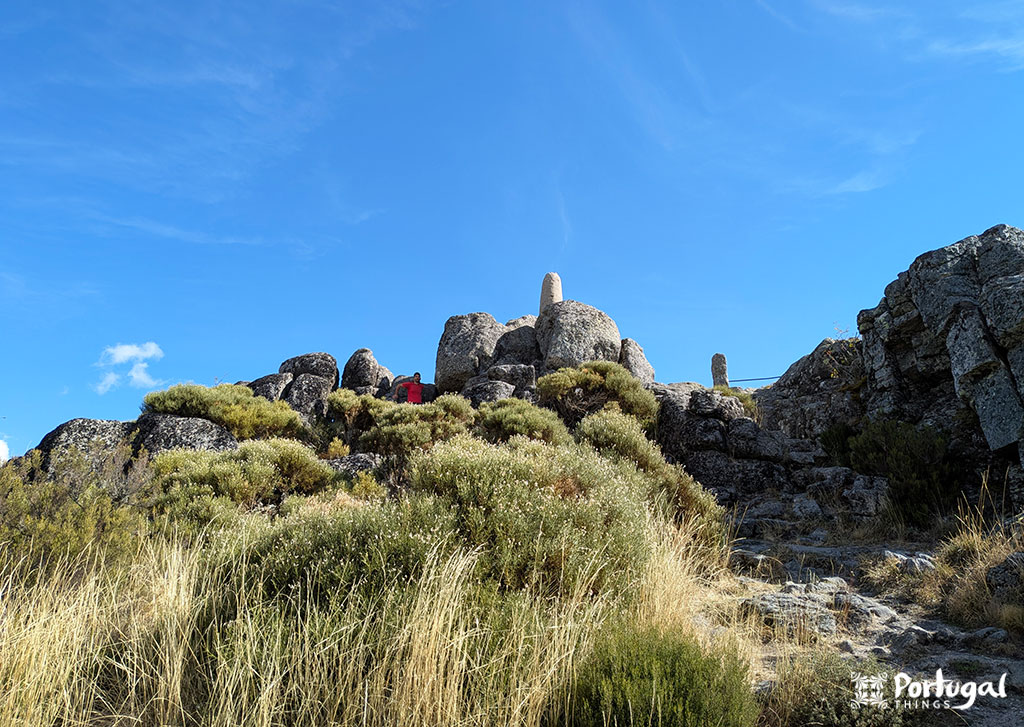 Rocky hill with large boulders and scattered dry grass under a light blue sky in Serra da Estrela. Two stone markers sit atop the hill, and a small red structure appears among the rocks. The Portugal logo is in the bottom right corner. Perfect for an easy trail adventure.
