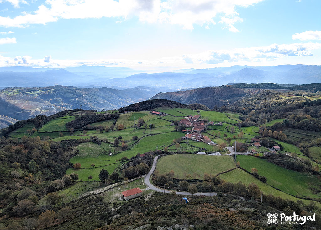 Uma pequena vila rural com campos verdes e casas espalhadas fica entre colinas e vales sob um céu parcialmente nublado, com montanhas distantes ao fundo. 