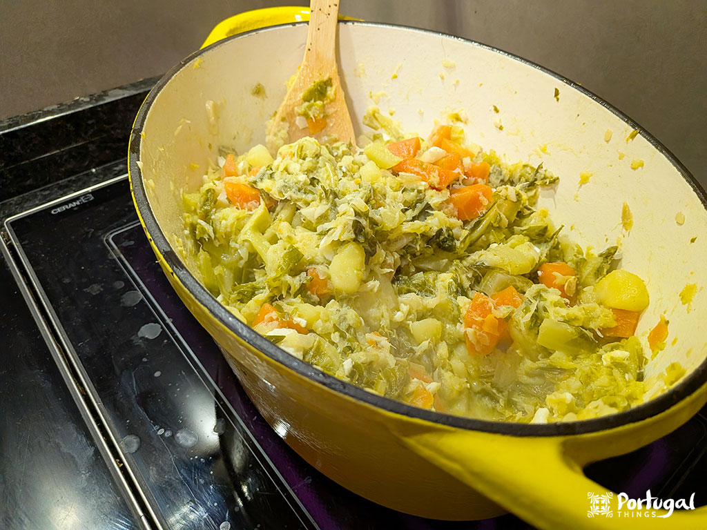 A yellow pot on the stove holds a mixture of chopped vegetables, including carrots, potatoes, and greens, being stirred with a wooden spoon. Steam rises as this Christmas recipe resembles the classic Roupa Velha.
