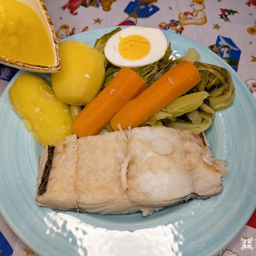 Um prato com bacalhau cozido, duas batatas, cenouras, folhas verdes e um ovo cozido meio clássico Bacalhau com todos. Uma mão derrama azeite de oliva amarelo por cima. A toalha de mesa festiva sugere essa tradicional Receita de Bacalhau.