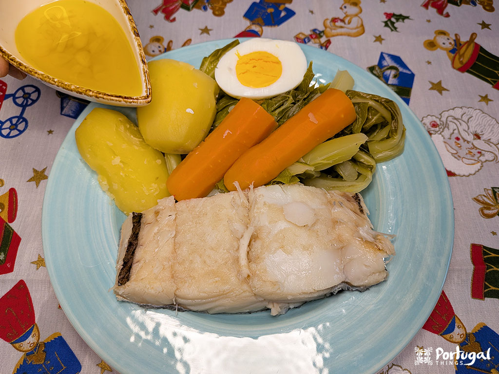A dish with boiled cod, two potatoes, carrots, leafy greens, and a boiled egg—classic bacalhau com todos. A hand drizzles golden olive oil over it. The festive tablecloth hints at this traditional codfish recipe.