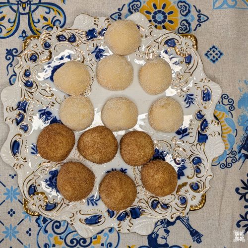 Um prato decorativo contém nove biscoitos Areias de Cascais revestidos de açúcar, seis de cor clara e três mais escuros, dispostos em um triângulo. O prato está apoiado em uma toalha de mesa azul e branca com tema português.