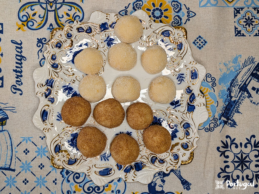 Um prato decorativo contém nove biscoitos Areias de Cascais revestidos de açúcar, seis de cor clara e três mais escuros, dispostos em um triângulo. O prato está apoiado em uma toalha de mesa azul e branca com tema português.