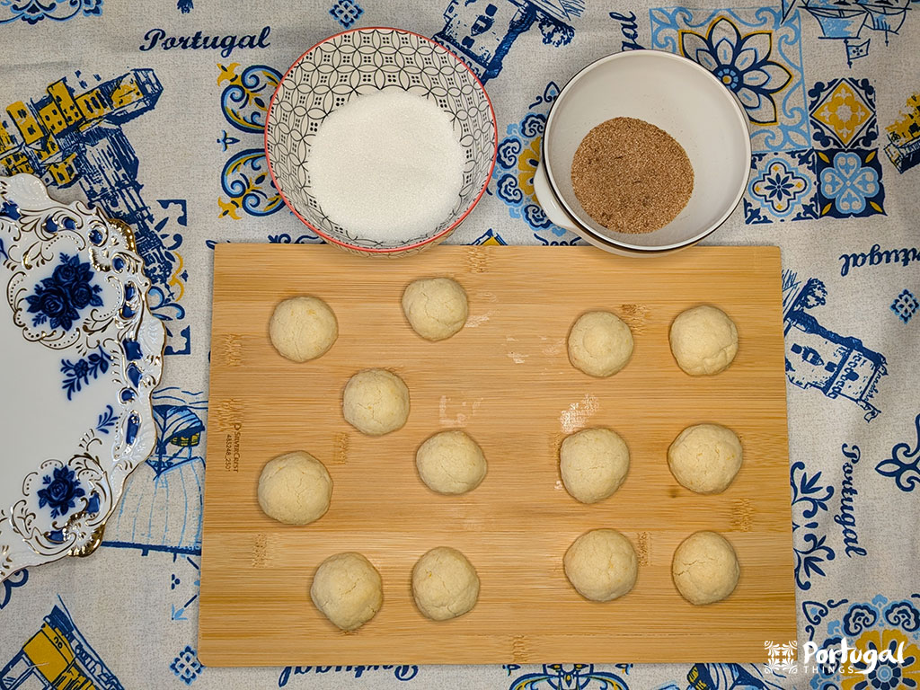 Uma tábua de madeira com 13 bolas de massa de Areias de Cascais ainda não assadas, uma tigela de açúcar e uma tigela de açúcar com canela em uma mesa coberta com um pano decorativo com tema português e pratos azuis e brancos - perfeito para uma receita fácil.
