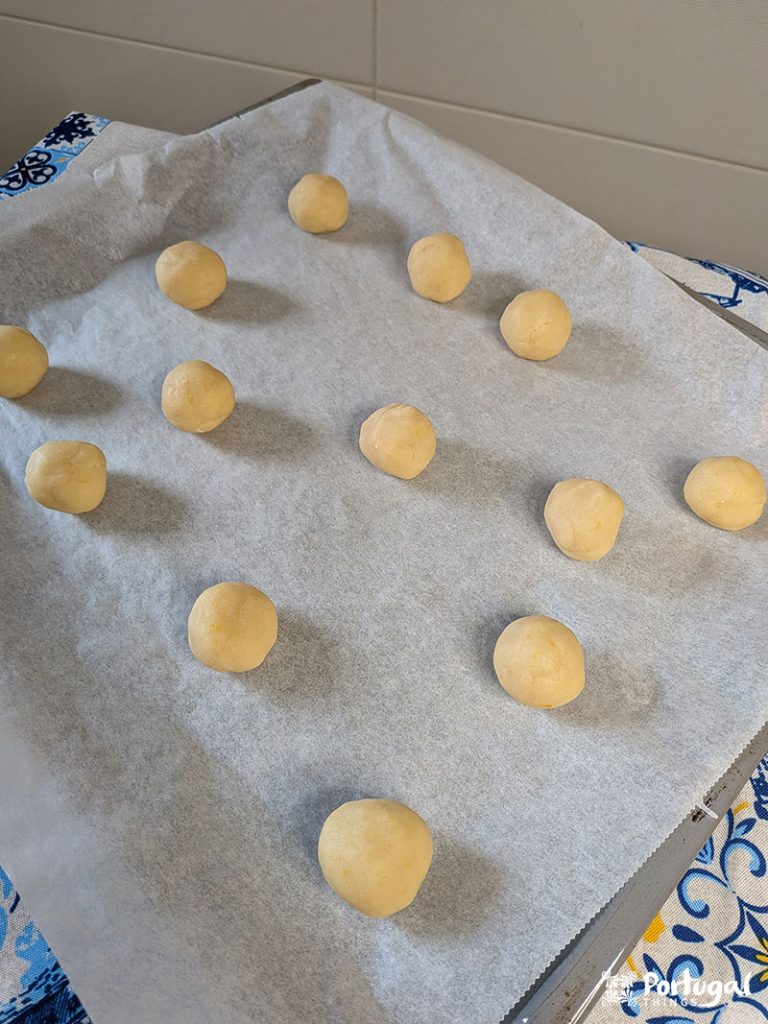 Bolas de massa de biscoito, inspiradas nas Areias de Cascais, estão uniformemente espaçadas em uma assadeira forrada de pergaminho, prontas para serem assadas. A bandeja repousa sobre uma superfície azul e branca estampada, prometendo um mimo Receita Fácil.