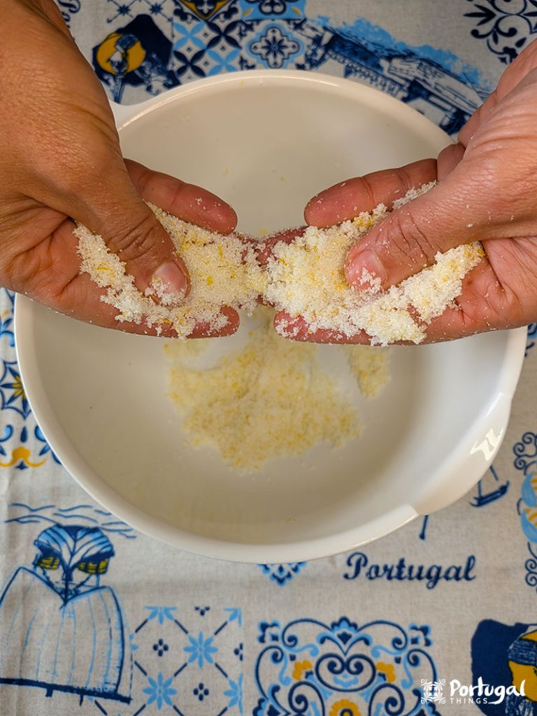 Duas mãos esfregam fubá sobre uma tigela branca, preparando a Receita Fácil para a Areias de Cascais. Grãos amarelos caem sobre uma toalha de mesa azul e branca.