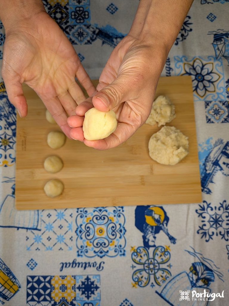 Uma pessoa molda a massa com as mãos sobre uma tábua de madeira com bolas de massa em uma toalha de mesa com estampa azul e amarela. 